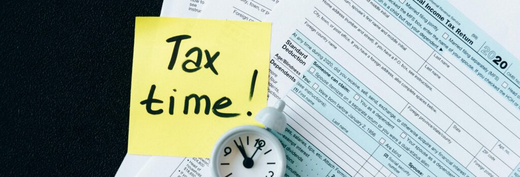 A stack of tax forms with a clock and yellow sticky note saying 'Tax time!' indicating urgency.